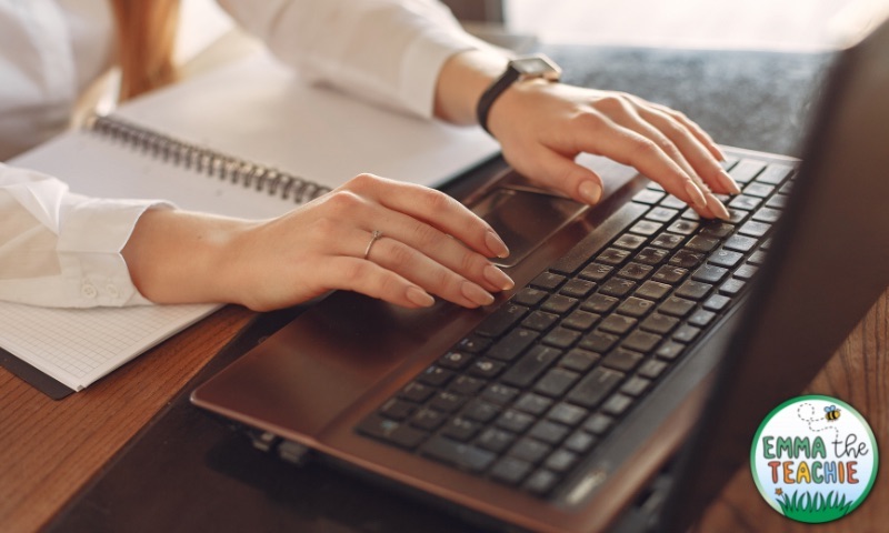 A woman wearing a white shirt types on a laptop, with a notepad on the desk in front of her.