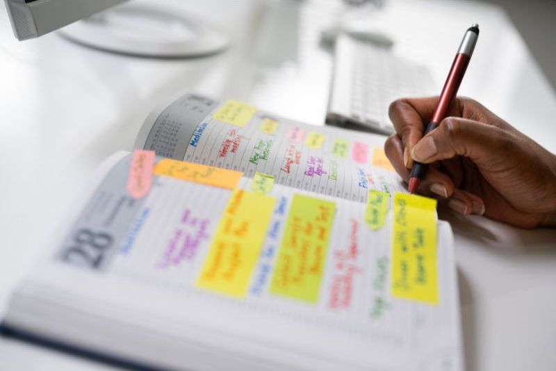 An image of a hand writing in an agenda. The agenda is full of yellow page markers with different times of events.