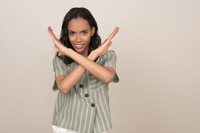 An image of a woman standing in front of a blank wall with her hands crossed in the shape of an X in front of her.