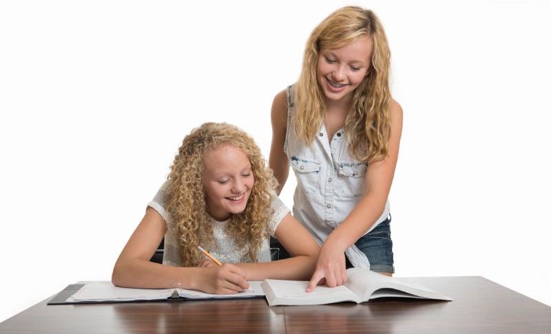 An image of two girls looking at a workbook on a table - one is pointing out a piece of the text, while the other is writing it down. Working together to peer review work is one way to cut down grading in science.