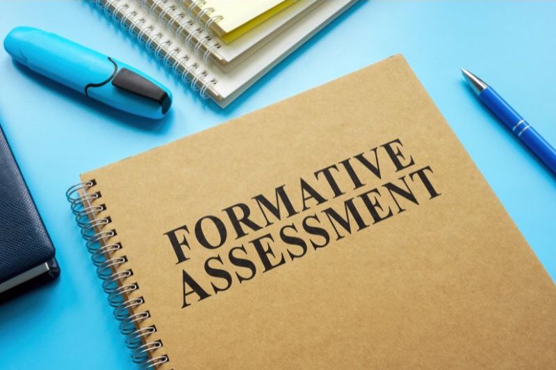 An image of a brown notebook with “Formative Assessment” written on the front. There is a blue highlighter, a blue pen, and a stack of notebooks surrounding the notebook. Using formative assessment is one way to cut down grading in science.