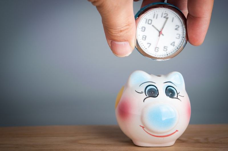 An image of a hand holding a mini clock. The mini clock is going into a mini piggy bank, symbolizing the concept of saving time.